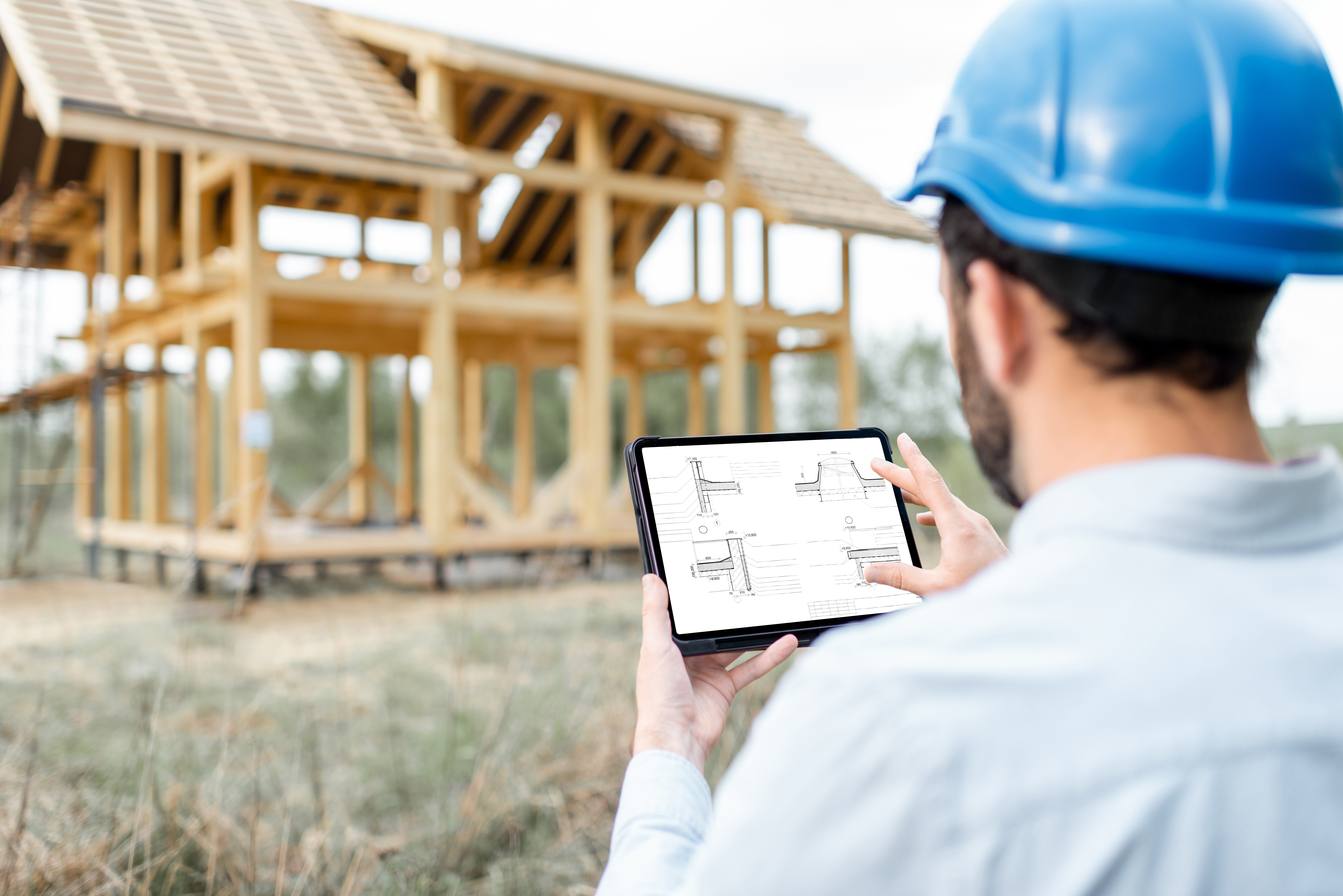 Engineer in a hard hat reviewing construction drawings on a tablet in front of a timber-framed house under construction
